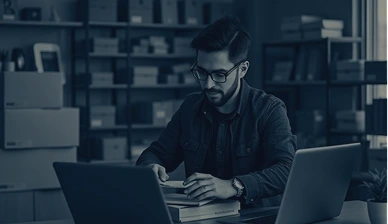 Person working at desk with laptops, books, and papers